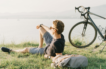 Young man with backpack and bicycle resting on summer meadow and enjoying view of sea.の写真素材