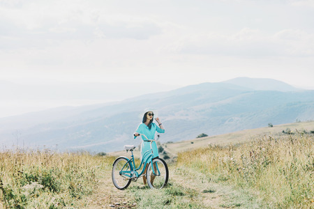Beautiful young woman wearing in dress and hat walking with bicycle on summer meadow.の写真素材