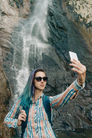 Traveler young woman with backpack taking selfie with smartphone on background of waterfall in summer.の写真素材