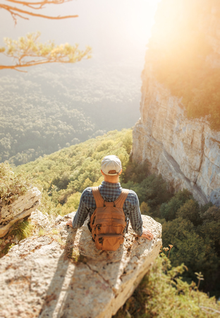Traveler young man with backpack sitting on edge of cliff over mountain valley in summer, rear view.の写真素材