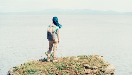 Backpacker young woman standing on peak of mountain and enjoying view of sea in summer, rear view.の写真素材