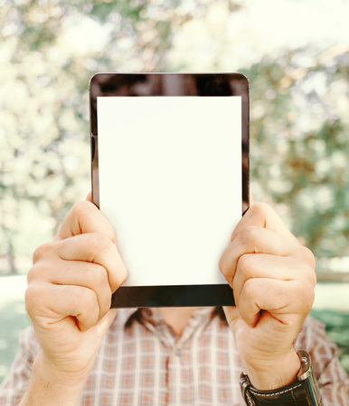 Young man holding tablet pc with empty screen in front of his face in summer park, close-up.の写真素材