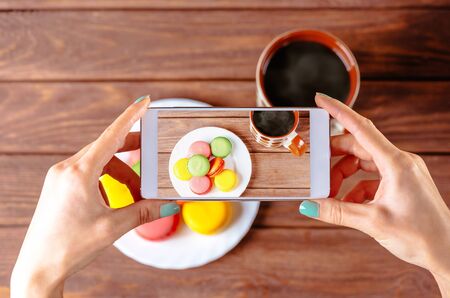 Woman taking photo of macarons dessert with cup of coffee with smartphone, top view of hands.の写真素材