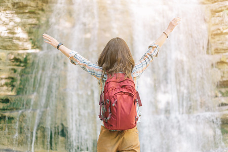Happy backpacker explorer young woman standing with raised arms in front of waterfall on sunny summer day outdoor, rear view.の写真素材