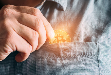 Male hand putting into pocket of shirt glowing gold bitcoin, symbol of crypto currency and virtual money.の写真素材