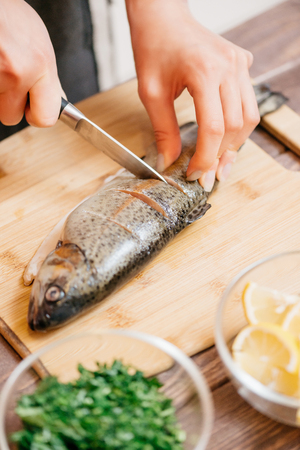 Woman cutting raw trout fish on wooden board for seafood dish in the kitchen, view of hands.の写真素材