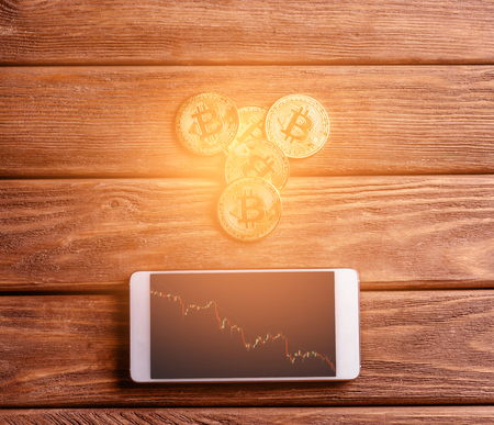 Many glowing gold bitcoins and smartphone with infographics on a wooden table.の写真素材