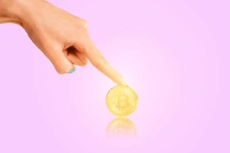 Female hand pointing to glowing gold coin bitcoin on a pink background.の写真素材