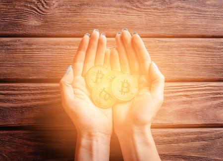 Many glowing gold bitcoins in female hands on a wooden background, top view.の写真素材
