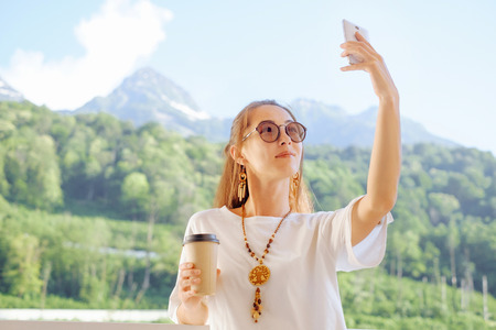 Smiling young woman with cup of coffee taking selfie with smartphone on balcony on background of...
