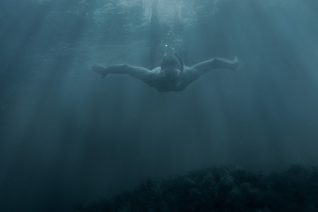 Active male freediver in mask swimming underwater among sunbeams, front view.
