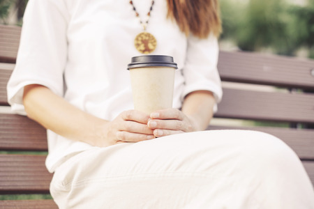Unrecognizable woman wearing in white clothing sitting with takeaway paper cup of coffee on wooden bench.の写真素材