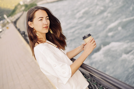 Smiling beautiful young woman walking with takeaway paper cup of coffee on embankment of river in...