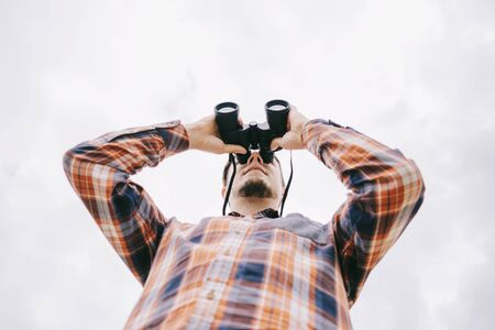 Bottom view of young man in plaid shirt watching with binoculars and exploring surroundings on white background of sky. Travel concept.の写真素材