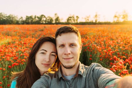 Smiling young loving couple taking photo selfie in red poppy flower meadow on sunny summer day, point of view.の写真素材