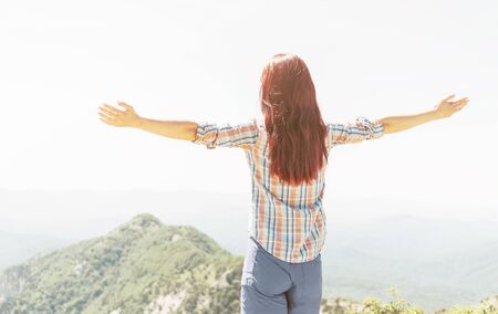 Rear view of freedom explorer young woman standing with raised arms in summer mountains.の写真素材