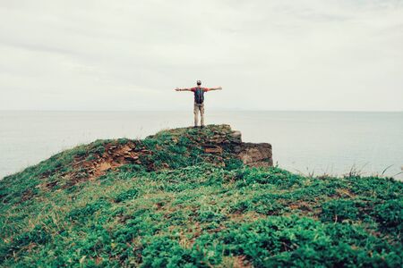 Happy hiker young man standing  with raised arms on peak of rock and enjoying view of seaの写真素材