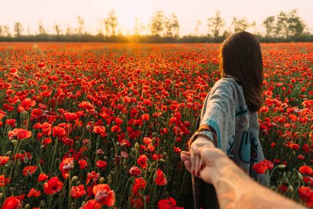 Couple in love. Man follows a brunette young woman in poppy flower meadow at summer sunset, point of view.の写真素材