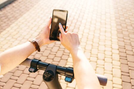Unrecognizable young woman using smartphone while standing with electric scooter in street outdoor, point of view in first person.の写真素材