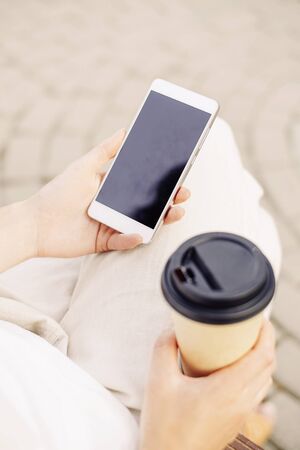 Unrecognizable young woman sitting with takeaway paper cup of coffee and smartphone, mock-up.の写真素材