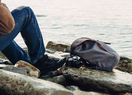 Traveler resting on coastline. Image of male legs, backpack and vintage photo camera on background of seaの写真素材