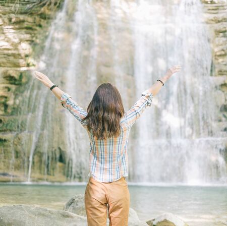 Happy explorer young woman standing with raised arms in front of waterfall.の写真素材