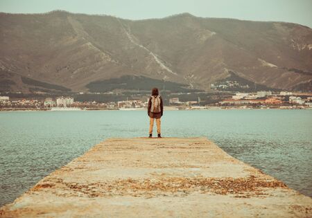 Unrecognizable traveler with backpack standing on pier at bay in front of mountains, rear viewの写真素材