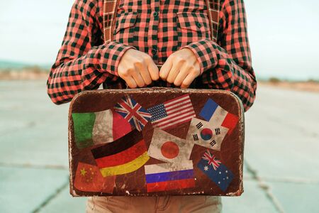 Woman holds small retro suitcase on road, face is not visible. Suitcase with stamps flags representing each country traveled.の写真素材