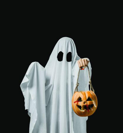 White ghost holding trick-or-treat pumpkin basket with sweet candy on dark background in Halloween holiday.の写真素材
