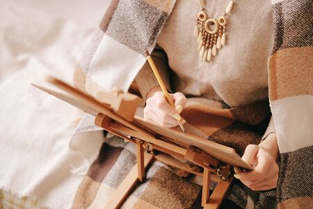 Young woman artist wrapped in plaid drawing a sketch on canvas easel with pencil indoor.の写真素材