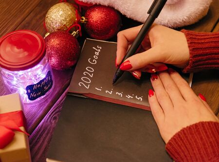 View of female hands writing 2020 goals in notebook on wooden table near New Year decorations.