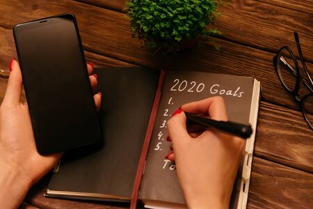 Woman writing 2020 goals in paper notepad and holding phone at work wooden table near plant and glasses, view of hands. Point of view.の写真素材