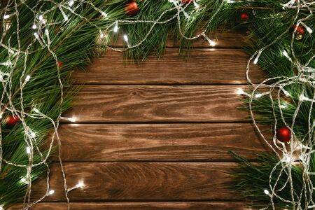 Christmas and New Year wooden background with fir-tree branches, red balls and led light garland decorations.