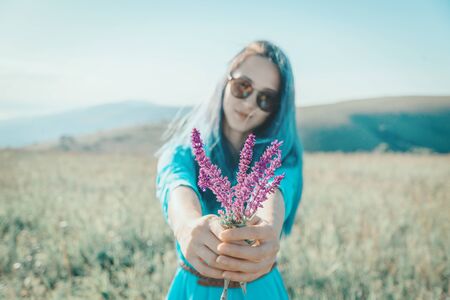 Stylish beautiful young woman with blue hair in dress and sunglasses giving bouquet of wildflowers and looking at camera in summer green grass meadow.の写真素材