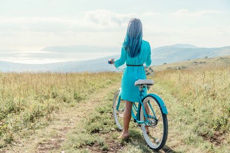 Girl with blue hair and wearing in dress standing with bicycle on country road along green meadows...