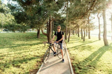 Casual style young man in sunglasses standing with bicycle on path in pine woods summer park.の写真素材