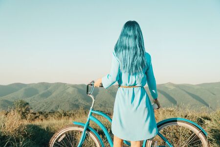 Stylish woman with blue hair standing with bicycle in front of mountain ridge in summer outdoor,...
