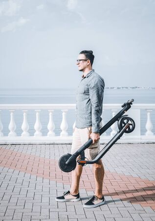 Casual style young man standing with folding electric scooter on city embankment in front of sea in summer.の写真素材