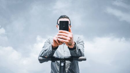 Young man standing with an electric scooter and using smartphone in front of cloudy sky, low angle...