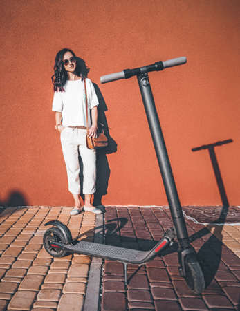 Fashionable beautiful young woman in white clothes standing near the wall, electric scooter in foreground.の写真素材
