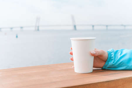 A glass of coffee on the background of the sea. Close-up of a female hand with a hot drink.の写真素材