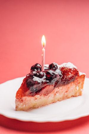 Delicious piece of berry cake with burning candles for a holiday on a pink background.