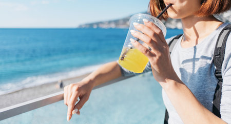 Beautiful young woman smiles and drinks fresh juice in summer in the background the sea. Focus on hand.の写真素材