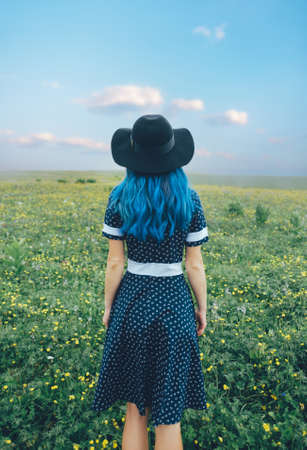 Woman in a hat and dress stands on a flower meadow on a clear summer day.