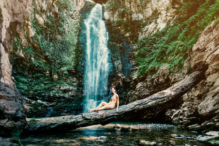 Young attractive woman sits on a tree on the background of a waterfall.の写真素材