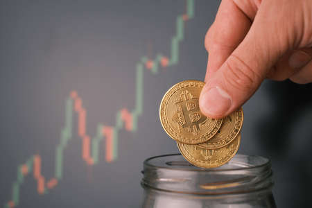 Man tosses a few bitcoin into a glass jar or piggy bank.