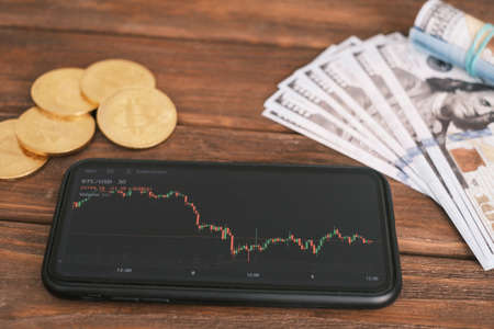 Phone with a graph, Bitcoin and Dollars lie on a wooden table.の写真素材