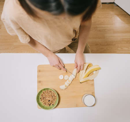Young woman slices breakfast banana with granola and yogurt. View from above.