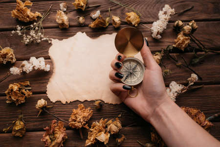 Woman hand holding compass in front of blank old paper among dry flowers.
