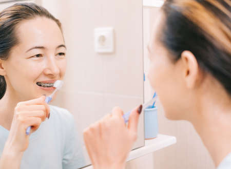 Young woman with braces cleans her teeth with a toothbrush, before mirror. Selective focus on the mirror.の写真素材